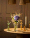 Der Überraschungs-Blumenstrauß mit lilafarbener Hortensie, zarten Gräsern und kleinen pinken Blumen steht in einer Glasvase auf einem runden Holztisch - ein charmantes Geschenk im warmen Licht.