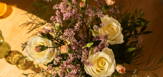 Ein Strauß aus weißen Rosen, kleinen rosafarbenen Blumen und zartem Grün, arrangiert in einer Vase, gebadet in warmem Sonnenlicht auf einer hölzernen Oberfläche.