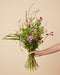 Eine Hand hält den handgebundenen Blumenstrauß Feenzauber in der Größe Classic mit rosa Bartnelken, dunkelrotem Wiesenknopf und filigranen Gräsern vor warmem beigem Hintergrund.