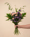 Eine Hand hält den Strauss So-kunterbunt-wie-du mit orangen Chrysanthemen, rosa Nelken, lila Lisianthus und lavendelfarbenem Statice vor einem warmen beigen Hintergrund.