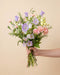 Eine Hand hält den Blumenstrauß 'Sommersorbet' in der Größe Classic mit violetten und rosa Sommerblumen vor beigem Hintergrund.
