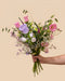 Eine Hand hält den Blumenstrauß 'Mariposa' in der Größe Classic mit rosa und violetten Blüten vor beigem Hintergrund.