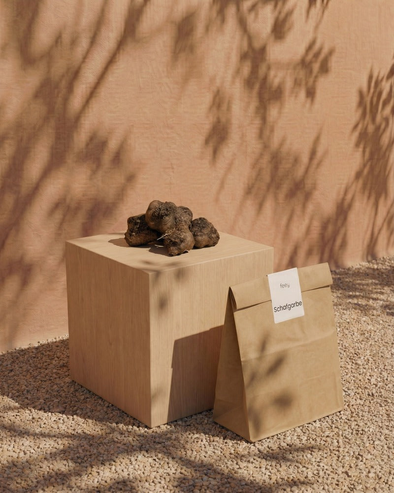 Schafgarbe-Rhizome und feey-Papiertüte auf einem Holzwürfel im warmen Sonnenlicht