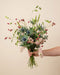Eine Hand hält den lockeren Wildblumenstrauß Bergluft mit blauen Edeldisteln und rosa Levkojen vor beigem Hintergrund