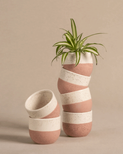 Drei Keramik Baby-Töpfe aus dem Set „Variado“ sind kreativ gestapelt und zeigen ihr gesprenkeltes Design mit rosa und cremefarbenen Streifen. Ein Topf beherbergt eine grüne Pflanze, während zwei andere kunstvoll daran gelehnt sind. Die Szene ist vor einem neutralen beigen Hintergrund dargestellt.