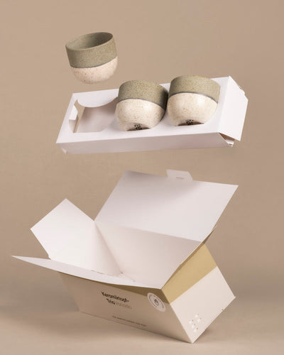 Ein Set des „Variado“-Keramik-Baby-Topfsets (9 Stk.) mit gesprenkeltem Beige- und Grün-Design schwebt über einer offenen weißen Pappschachtel vor einem schlichten beigen Hintergrund und erinnert an die Lieblingsfarbe, perfekt für die Pflege Ihrer Babypflanzen-Träume.