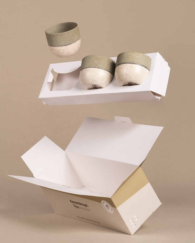 Drei Keramik-Baby-Töpfe aus dem Set „Variado“ in grün-beigem Design hängen über einer offenen, minimalistischen weißen Kartonschachtel vor beigem Hintergrund. Die Verpackung ist mit Branding und Umweltsymbolen versehen.