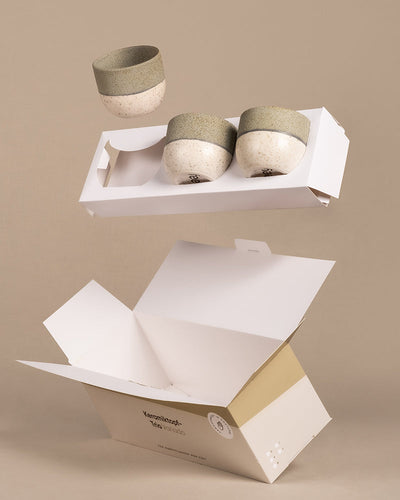 Drei Keramik-Baby-Töpfe aus dem Set „Variado“ in grün-beigem Design hängen über einer offenen, minimalistischen weißen Kartonschachtel vor beigem Hintergrund. Die Verpackung ist mit Branding und Umweltsymbolen versehen.