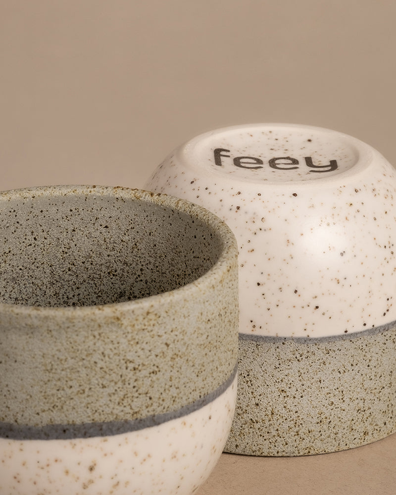 Zwei Keramiktassen aus dem Set „Variado“ mit einem gesprenkelten Design in neutralen Tönen, das an elegante Keramiktöpfe erinnert. Eine Tasse ist aufrecht dargestellt, sodass die Innenseite sichtbar ist, während die andere umgedreht ist und auf ihrem Boden den Markennamen „feey“ trägt. Der schlichte beige Hintergrund unterstreicht dieses harmonische Arrangement.