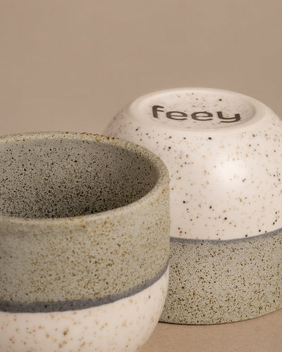 Zwei Keramiktassen aus dem Set „Variado“ mit einem gesprenkelten Design in neutralen Tönen, das an elegante Keramiktöpfe erinnert. Eine Tasse ist aufrecht dargestellt, sodass die Innenseite sichtbar ist, während die andere umgedreht ist und auf ihrem Boden den Markennamen „feey“ trägt. Der schlichte beige Hintergrund unterstreicht dieses harmonische Arrangement.