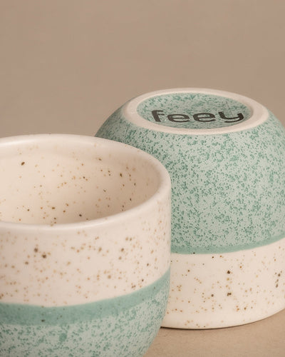 Zwei Tassen aus dem Keramik Baby-Topfset 'Variado' (6 Stk.) weisen ein gesprenkeltes Design auf, das sanftes Türkis mit weißen Farbtönen verbindet und an raffinierte deutsche Handwerkskunst erinnert. Eine Tasse steht aufrecht, während die andere umgedreht ist. Das charmante gepunktete Muster zeigt auf einer Unterseite „Feey“ und kommt vor einem neutralen beigen Hintergrund wunderschön zur Geltung.