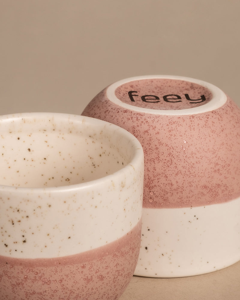 Zwei Keramiktassen mit gesprenkeltem Design, die an das entzückende Keramik-Baby-Topfset „Variado“ erinnern. Eine Tasse liegt auf der Seite, sodass auf der Unterseite das Wort „feey“ zu sehen ist. Jede Tasse hat in der Mitte ein rosa Band, das mit einem creme- und braunfarbenen gesprenkelten Muster verziert ist.