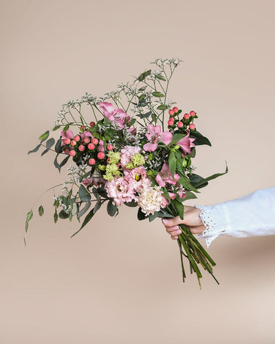 Eine Hand hält den Ballerina-Strauß, der aus verschiedenen Blumen mit rosafarbenen Rosa-Blüten, roten Beeren, grünen Blättern und zarten weißen Füllungen besteht und vor einem schlichten beigen Hintergrund steht.