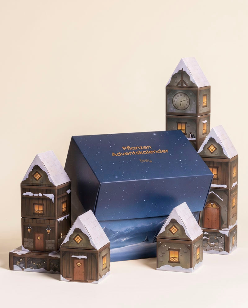Geschlossene blaue Adventskalender-Box, flankiert vom verschneiten Papierhäuser-Winterdorf mit Uhrturm vor beigem Hintergrund.