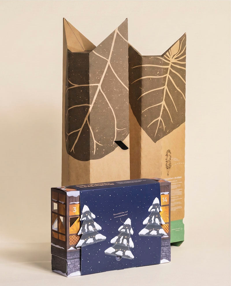 Aufgeklappte Kraftpapier-Hülle mit Blattmotiv hinter der blauen Adventskalender-Box mit Tannenmotiv vor beigem Hintergrund.