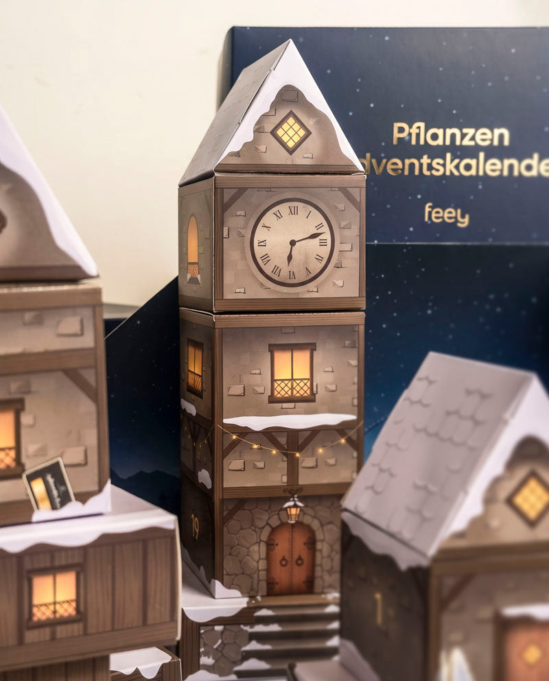 Nahaufnahme des beleuchteten Uhrturm-Häuschens im Adventskalender-Winterdorf Abendblau mit verschneiten Dächern und der feey-Box im Hintergrund.
