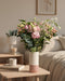 Der Blumenstrauß Ballerina mit rosa und cremefarbenen Blüten und Eukalyptus steht in einer zweifarbigen Keramikvase auf einem Holztisch neben einer Tasse und Büchern; im Hintergrund ein Bett mit hellen Kissen.