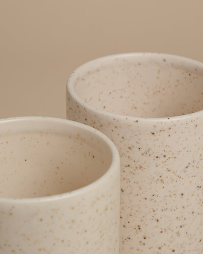 Zwei weiße Keramikbecher mit gesprenkeltem Muster stehen nebeneinander auf einem beigen Hintergrund. Ihre glatten, abgerundeten Kanten und das minimalistische Design passen perfekt zur feey Keramik-Vase (Variado | 21 cm).