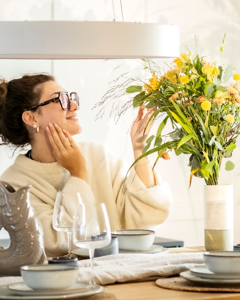 Eine Frau mit Brille und cremefarbenem Pullover lächelt, während sie einen gelben Blumenstrauß in der feey Keramik-Vase (Variado | 21 cm) bewundert, die auf einem gedeckten Esstisch mit Tellern, Gläsern und einem fischförmigen Krug steht.