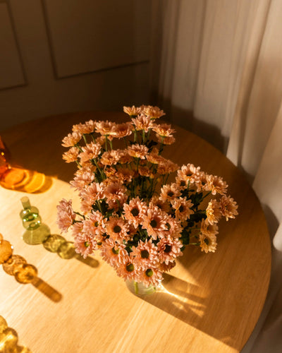 Chrysanthemen pfirsich (10 Stk.) in einer Glasvase stehen auf einem runden Holztisch im Sonnenlicht, schaffen eine blühende Atmosphäre mit Deko-Glasobjekten und transparenten Vorhängen im Hintergrund.