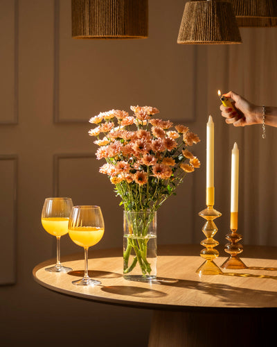 Eine Hand zündet eine Spitzkerze neben Chrysanthemen-Pfirsich (10 Stk.) und zwei Gläsern mit Orangengetränk auf einem runden Holztisch an und schafft eine blühende Atmosphäre unter warmem Licht und gewebten Lampenschirmen.