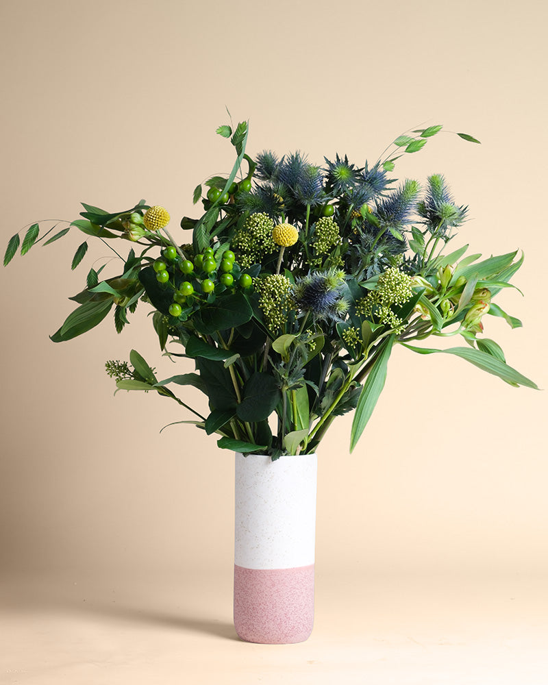 Die Erfolgsgeheimnis-Vase in Weiß und Rosa zeigt ein frisches Bouquet mit grünem Laub, runden gelben Blüten, grünen Beeren und blauen Disteln, das vor einem neutralen beigen Hintergrund positive Energie ausstrahlt.