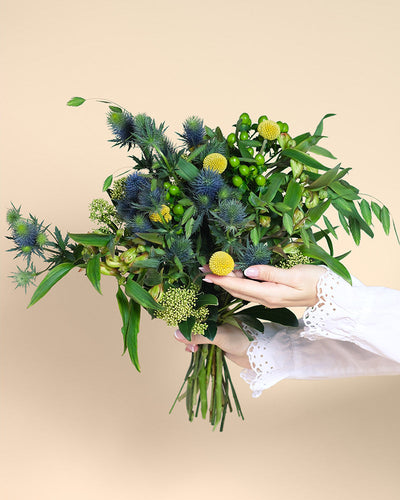 Eine Person in einer weißen Bluse mit gekräuselten Manschetten hält den Erfolgsgeheimnis-Strauß aus üppigem Grün, blauen Disteln und gelben Bällen, der vor einem schlichten beigen Hintergrund Ruhe ausstrahlt.