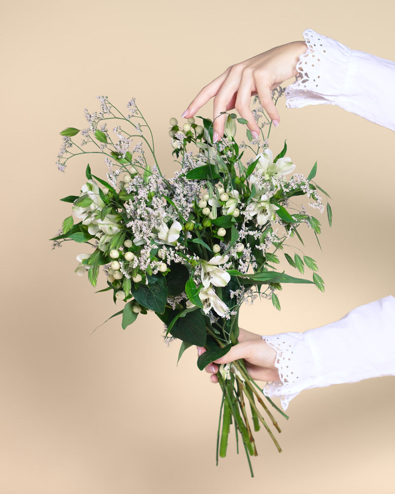 Eine Hand in einem weißen, mit Spitze besetzten Ärmel hält den Luftkuss-Strauß - grüne Blätter, weiße Blüten und kleine weiße Beeren - vor einem neutralen beigen Hintergrund.