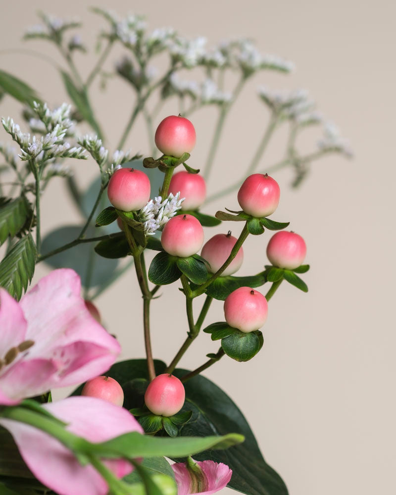 Nahaufnahme des Ballerina-Straußes mit rosafarbenen Hypericum-Beeren auf grünen Stielen, akzentuiert durch kleine weiße Blüten und eine teilweise sichtbare rosafarbene Lilie, alles vor einem weichen beigen Hintergrund.