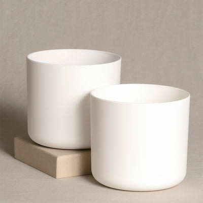 Zwei minimalistische, zylindrische, weiße Keramik-Topfsets (2 x 14) werden auf einem neutralen Hintergrund präsentiert. Eines ist etwas kleiner als das andere und sie stehen nebeneinander auf einem rechteckigen beigen Block. Die Pflanzgefäße haben eine glatte, matte Oberfläche, perfekt für die Präsentation von Zimmerpflanzen.