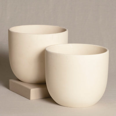 Zwei leichte Topfsets (2 x 16) in Beige stehen auf einem neutralen Hintergrund. Die Pflanzgefäße sind modern, zylindrisch geformt und haben eine glatte, matte Oberfläche. Der kleinere Topf steht leicht vor und links vom größeren und ruht auf einer kleinen quadratischen Fliese.