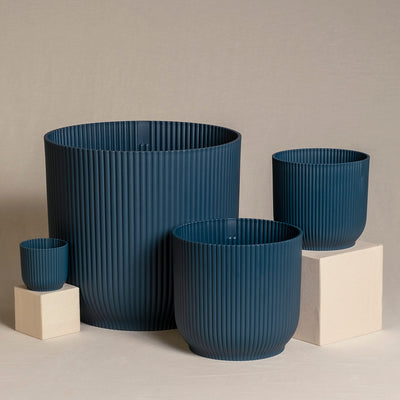 Ein Set aus vier blauen, gerippten Topfsets (30, 18, 14, 7) unterschiedlicher Größe, angeordnet auf neutralem Untergrund. Der kleinste Topf sitzt auf einem kleinen weißen Würfel, während ein anderer, etwas größerer Topf auf einem größeren weißen Würfel steht. Die anderen beiden Feey-Töpfe stehen direkt auf der Oberfläche.