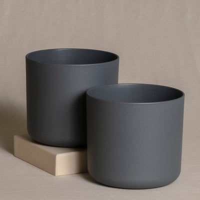 Zwei mattschwarze zylindrische Topfsets (2 x 14) unterschiedlicher Größe stehen auf einer beigen Fläche mit neutralem Hintergrund. Der kleinere Topf aus hochwertigem Kunststoff steht leicht erhöht auf einem kleinen beigen Block und verleiht so eine minimalistische und moderne Ästhetik für Zimmerpflanzen.