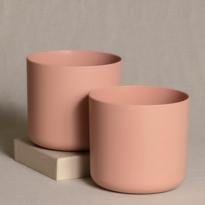 Zwei minimalistische, mattrosa Topfsets (2 x 14) werden vor einem neutralen beigen Hintergrund präsentiert. Ein Pflanzgefäß ist etwas höher und steht auf einem kleinen beigen Block, während das andere, niedrigere Pflanzgefäß davor positioniert ist, wodurch ein Schichteffekt entsteht – perfekt für die Präsentation von Zimmerpflanzen.