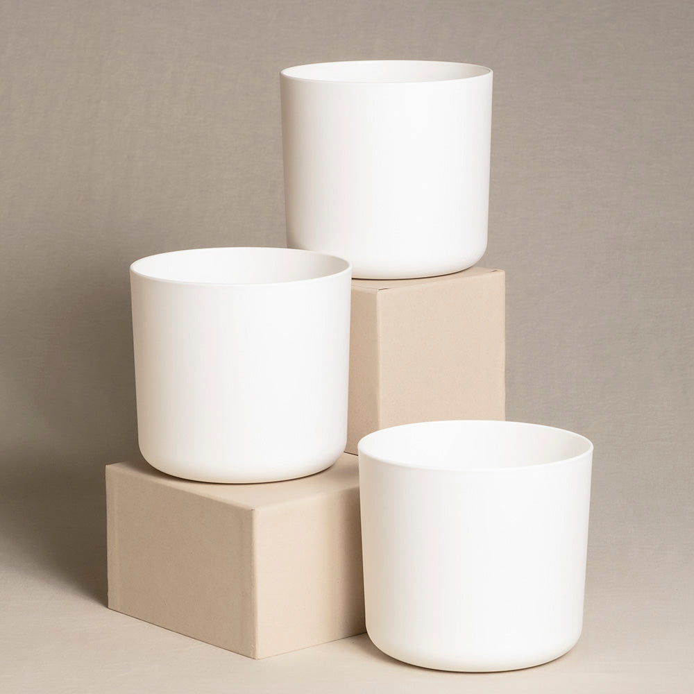 Drei weiße zylindrische Topfsets (3 x 16) in unterschiedlichen Höhen werden auf und um zwei beige rechteckige Blöcke vor einem neutralen Hintergrund ausgestellt. Das minimalistische Design betont die klaren Linien und glatten Oberflächen der Pflanzgefäße, perfekt für die Präsentation von Zimmerpflanzen in jedem modernen Raum.