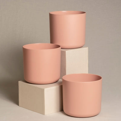 Drei minimalistische, mattrosa Topfsets (3 x 16) sind auf und um zwei gestapelte beige Würfel angeordnet. Ein Topf sitzt auf dem oberen Würfel, ein anderer auf einem unteren Würfel und der dritte direkt auf dem Boden. Der neutrale Hintergrund hält den Fokus auf diese stilvollen Blumentöpfe.