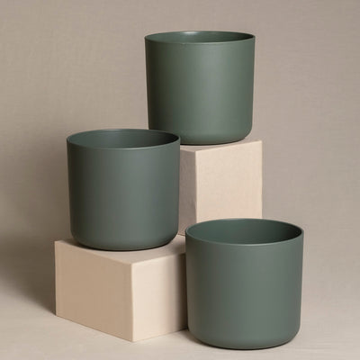 Drei dunkelgrüne Topfsets (3 x 16) stehen auf zwei beigen Blöcken, die in unterschiedlichen Höhen angeordnet sind. Sie stehen vor einem neutralen Hintergrund. Das minimalistische und moderne Design verleiht der Komposition ein elegantes und sauberes Aussehen.