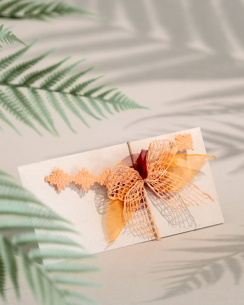 Ein Gutschein, der mit einer großen orangefarbenen Netzschleife und aufwendigen Banddetails verziert ist, liegt auf einer hellen Oberfläche. Schatten von Farnblättern, die über die Szene geworfen werden, erzeugen eine sanfte, natürliche Atmosphäre – ideal als bezauberndes Geschenk.