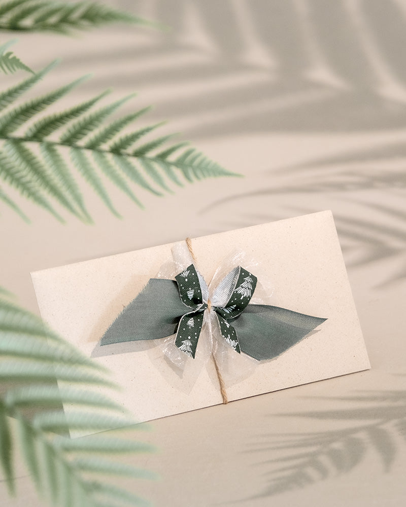 Ein Umschlag mit einem Gutschein, der mit einer graugrünen Schleife zusammengebunden ist, ruht elegant auf einem beigen Hintergrund. Farnblätter im Vordergrund werfen sanfte Schatten und schaffen eine ruhige Umgebung, die sich perfekt für die Präsentation dieses besonderen Geschenks eignet.