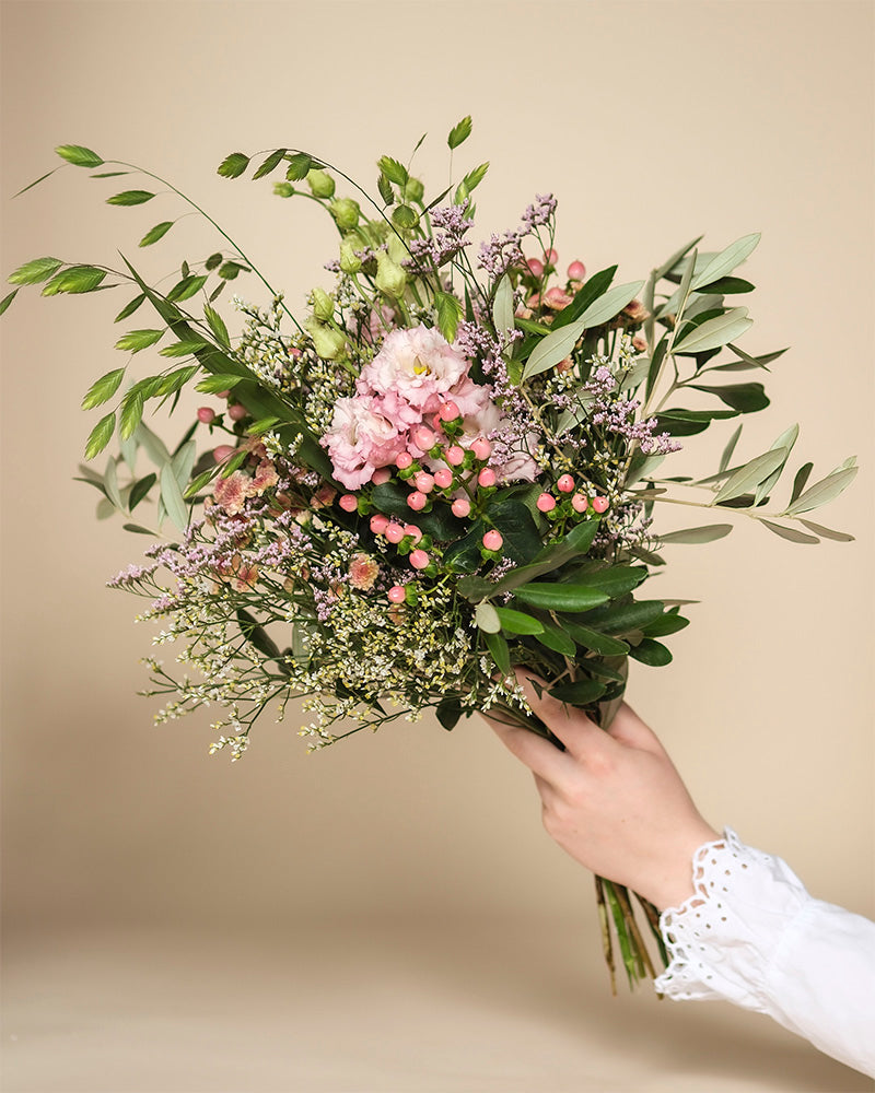 Eine Hand in einem weißen Spitzenärmel hält den Morgenlichtstrauß aus rosa Blumen, grünen Blättern und kleinen Herbstblüten vor einem schlichten beigen Hintergrund.