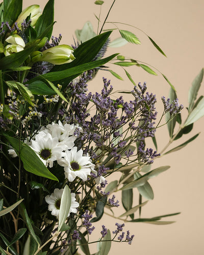 Der Blumenstrauß "Nebelmeer" verzaubert mit weißen Gänseblümchen, zarten Blüten, hellgrünen Knospen und purpurnem Statice auf beigem Hintergrund - wie ein Hauch Morgentau für jeden Anlass.
