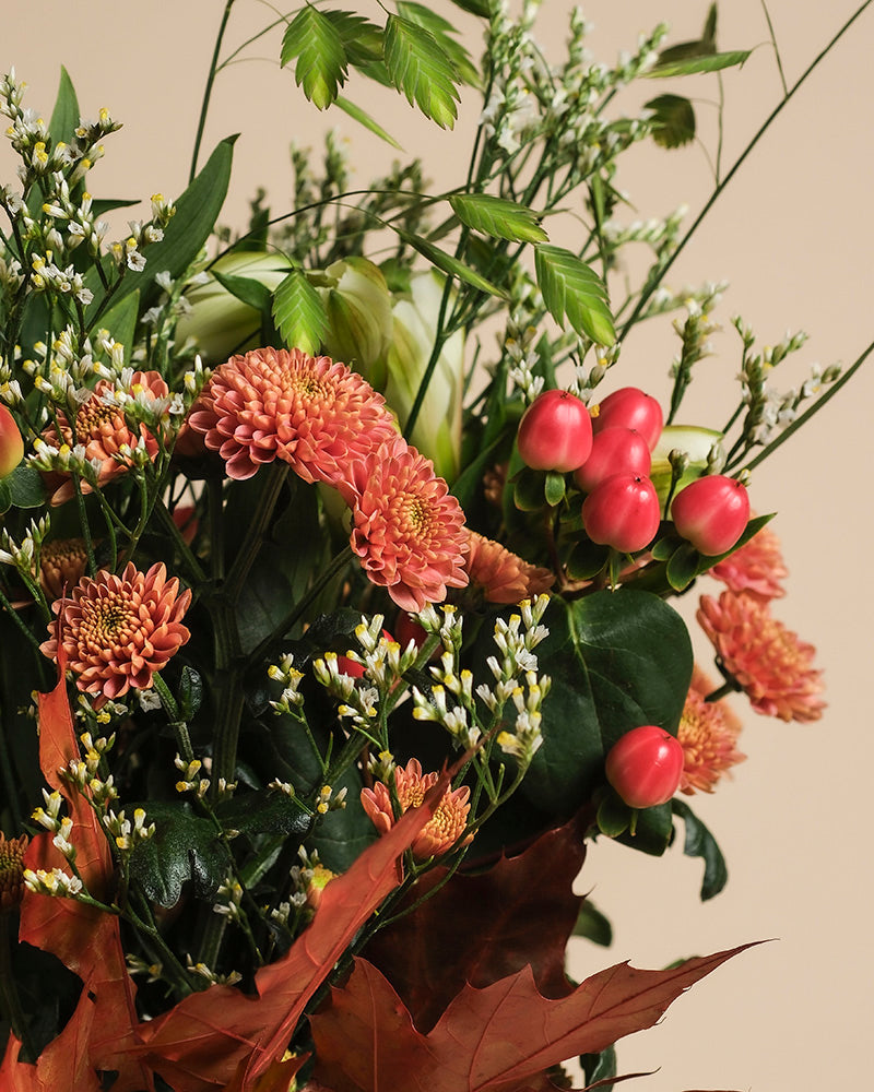 Herbstfreude, mit orangefarbenen Chrysanthemen, roten Beeren, kleinen weißen Blüten und grün-braunem Herbstlaub, schafft ein fröhliches Farbspiel vor einem beigen Hintergrund.