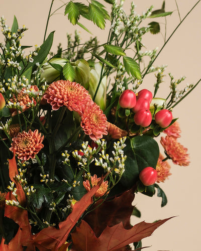 Herbstfreude, mit orangefarbenen Chrysanthemen, roten Beeren, kleinen weißen Blüten und grün-braunem Herbstlaub, schafft ein fröhliches Farbspiel vor einem beigen Hintergrund.