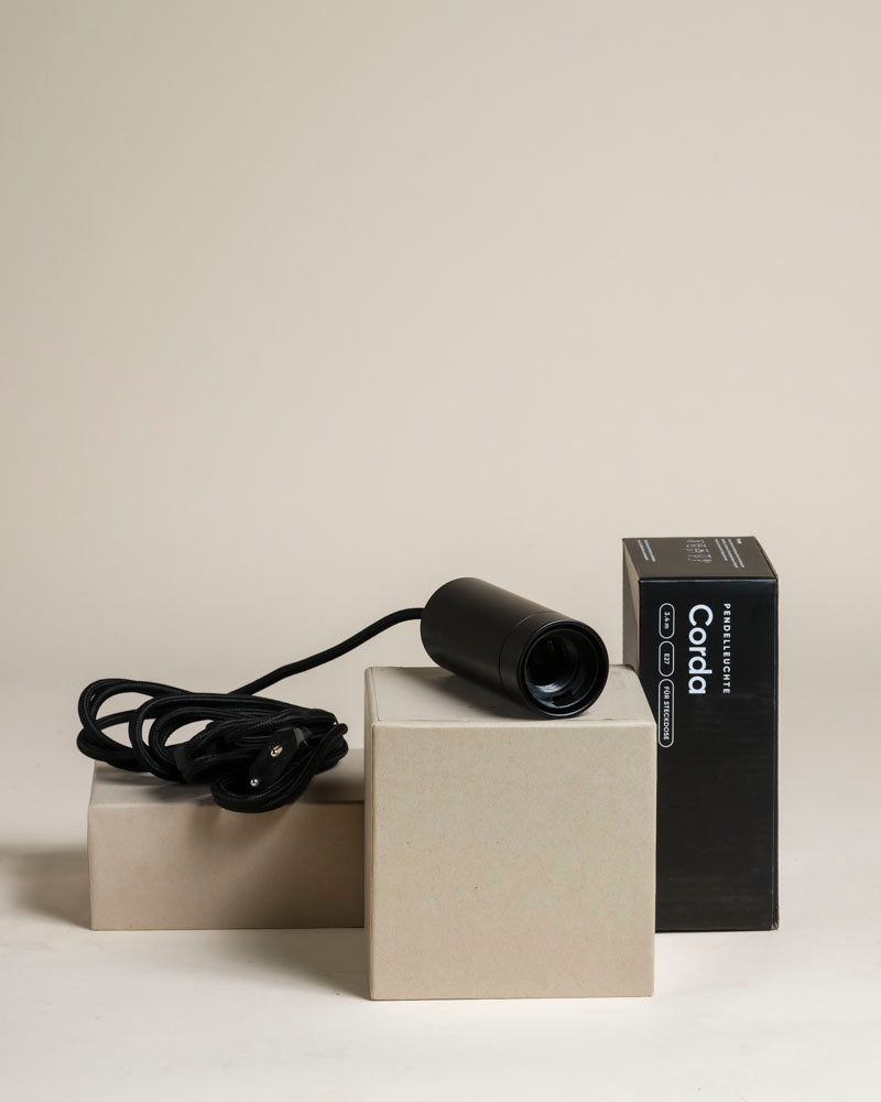 Eine schwarze Pendelleuchte 'Corda' mit Schnur sitzt auf beigen Blöcken neben ihrer schwarzen Box mit der Aufschrift "Corda" vor einem neutralen Hintergrund - ideal für die Beleuchtung von Zimmerpflanzen oder zur Aufwertung eines Raums.