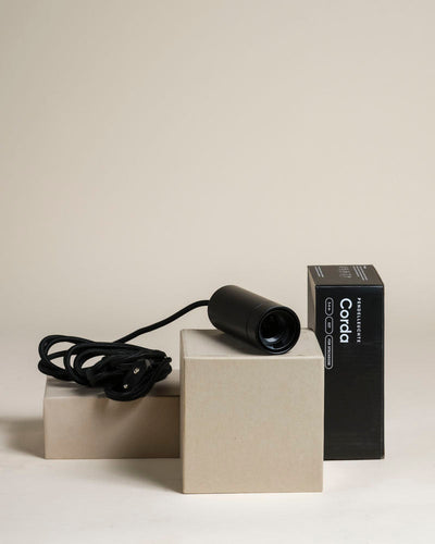 Eine schwarze Pendelleuchte 'Corda' mit Schnur sitzt auf beigen Blöcken neben ihrer schwarzen Box mit der Aufschrift "Corda" vor einem neutralen Hintergrund - ideal für die Beleuchtung von Zimmerpflanzen oder zur Aufwertung eines Raums.