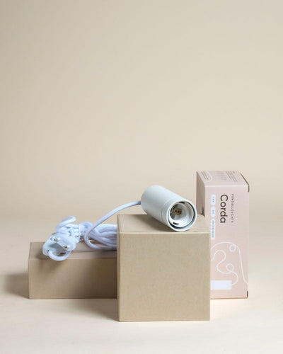 Die weiße Pendelleuchte 'Corda' mit Kordel und Stecker ist auf zwei gestapelten Kartons neben der beigen "Coda"-Verpackung abgebildet und eignet sich damit ideal zur Hervorhebung von Zimmerpflanzen vor einem schlichten beigen Hintergrund.