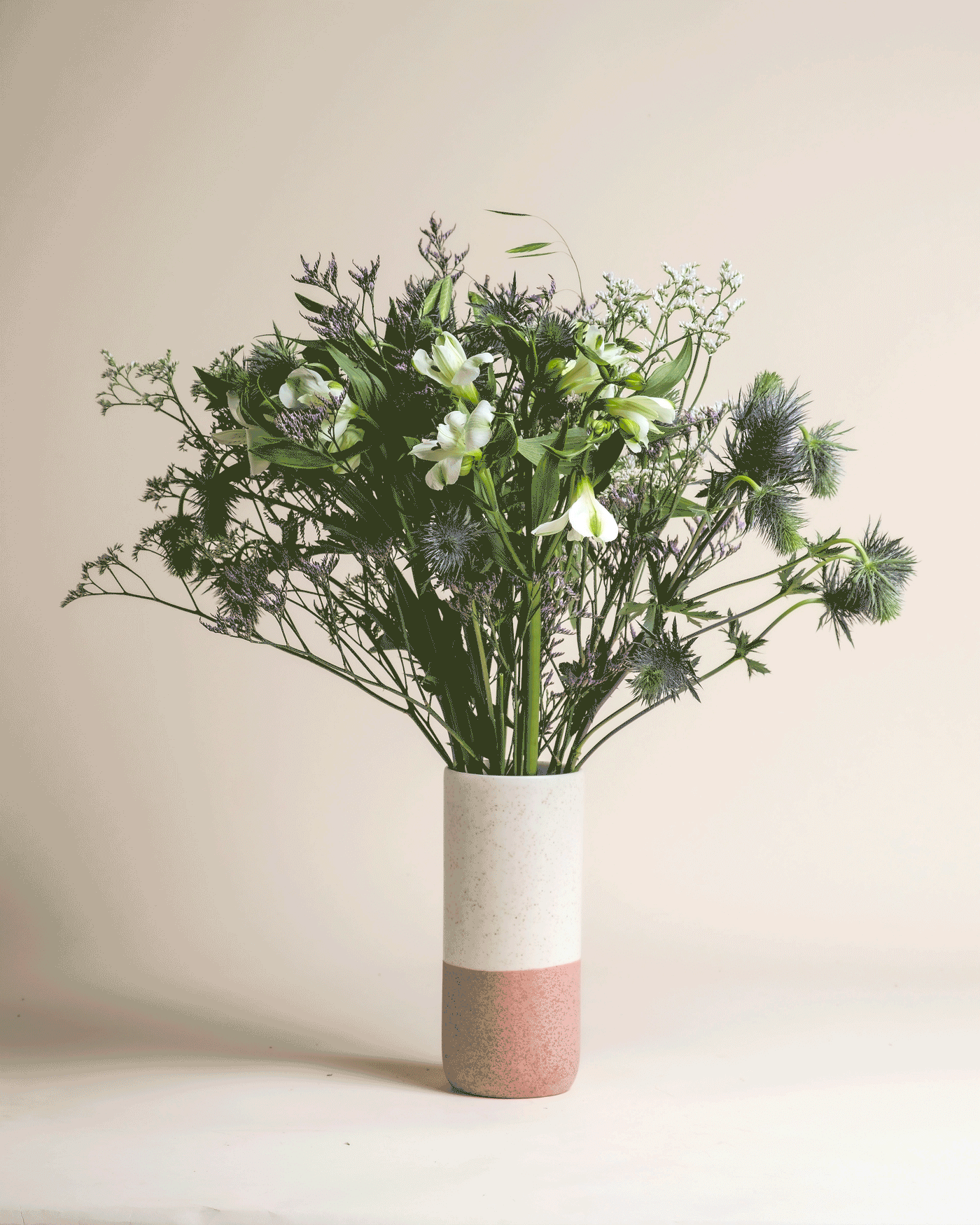 Die Keramikvase "Wanderlust" mit weißem und rosafarbenem Blockdesign enthält grüne und weiße Blumen, üppiges Grün und blaue Disteln, die an die Frische der Bergluft vor einem schlichten beigen Hintergrund erinnern.