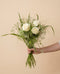 Eine Hand hält den Ich-denk-an-dich-Strauss mit weissen Rosen, zarten Trockenblumen und üppigem Grün vor einem warmen beigen Hintergrund.