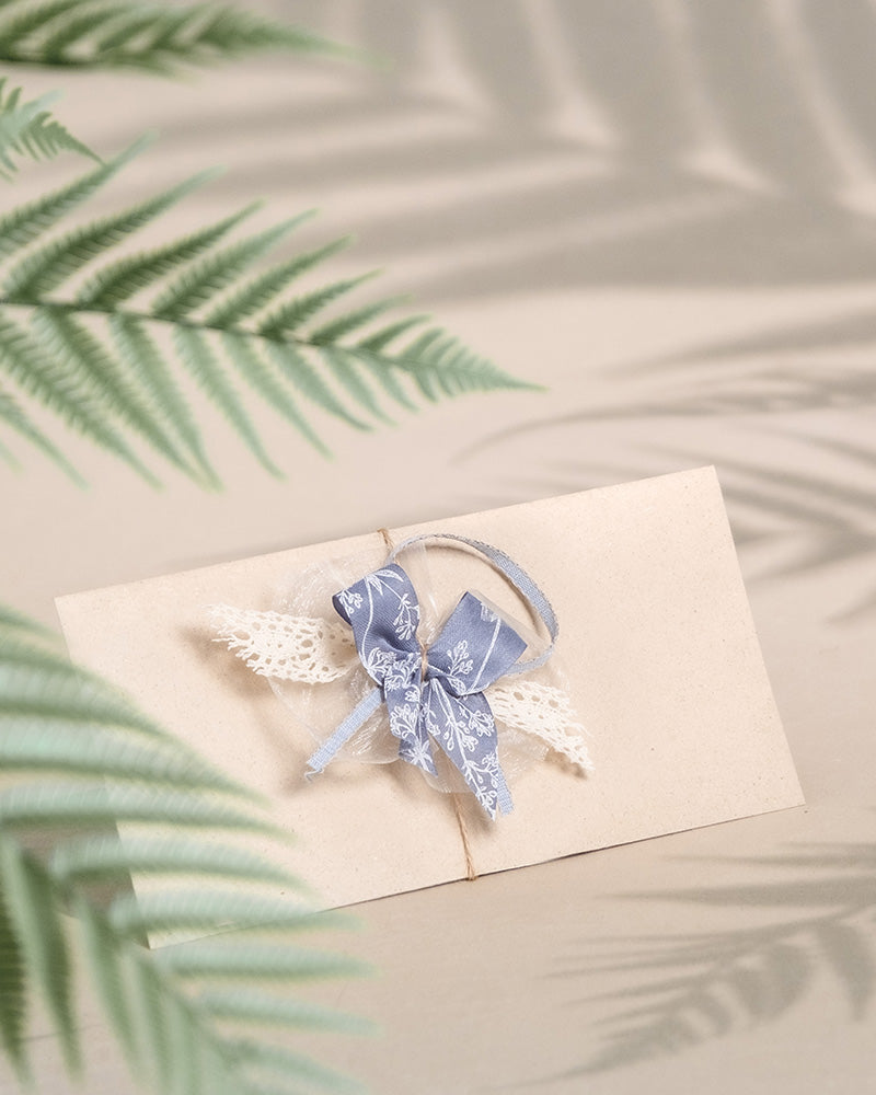 Ein beigefarbener, elegant mit einem blauen Band und Spitze verzierter Umschlag liegt auf einer hellen Oberfläche und erinnert an einen edlen Gutschein. Farnschatten erzeugen im Hintergrund ein zartes Muster und unterstreichen den natürlichen Charme des Umschlags.