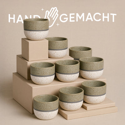 Acht Keramiktassen des Sets „Keramik Baby-Topfset Variado“ mit grünem Rand und gesprenkelter Oberfläche stehen auf beigefarbenen, gestapelten Boxen. Der beige Hintergrund ist mit dem Schriftzug „HANDGEMACHT“ und einer darüber abgebildeten Hand verziert.